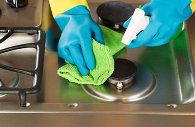دستانی با دستکش‌های دو رنگ آبی و زرد در حال اسپری کردن روی سطح گاز و پاک کردن آن با یک دستمال سبز 