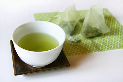 دو کیسه چای سبز بر روی یک دستمال سبز رنگ در کنار یک کاسه کوچک سفید حاوی مقداری چای سبز 