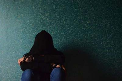 دختری با سوییت شرت مشکی و شلوار جین و دستبندهای مهرهای در حالی که نشسته و صورتش با کلاهش پوشانده شده