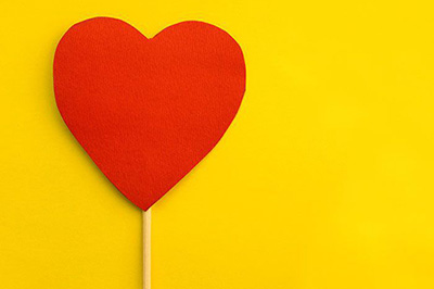 یک قلب قرمز کاغذی با دسته چوبی که بر روی یک صفحه زرد رنگ قرار دارد
