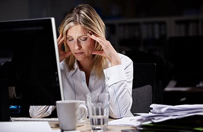 زن میانسال در حالیکه پشت کامپیوتر نشسته و چشمانش را بسته و دستانش را کنار شقیقه‌هایش گذاشته است