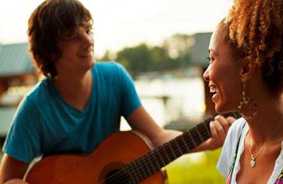 زن با پوست تیره و موهای قهوه‌ای در حالی که لبخند میزند به پسر که در حال گیتار زدن و خواندن است نگاه می‌کند