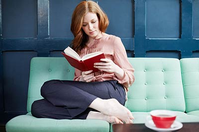 زن جوان با موهای قرمز در حالی که روی یک کاناپه آبی آسمانی نشسته و روی میز پیش رویش یک فنجان قرمز رنگ قرار دارد ، در حال خواندن یک کتاب با جلد قرمز است