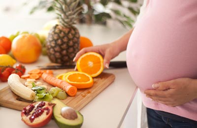 شکم یک زن باردار با لباس صورتی در کنار میزی پر از میوه های رنگارنگ مانند آناناس ، پرتقال، آووکادو
