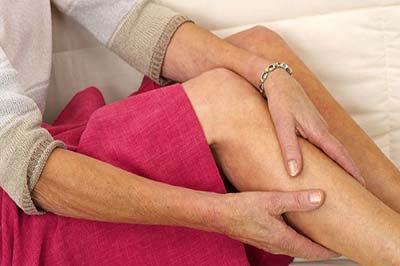 یک زن مسن با بلوز یاسی و دامن صورتی در حال ماساژ ساق پای راست خود