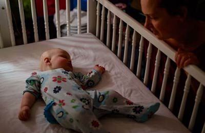 زنی کنار تخت نوزادش نشسته و به نوزاد که داخل تخت خوابیده نگاه می‌کند