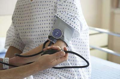 یک مرد با دستگاه فشار خون در حال اندازه گیری فشار خون زنی که لباس بیمارستان به تن دارد و روی تخت بیمارستان نشسته