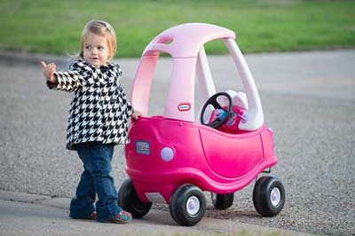 دختر خردسال زیبا در کنار ماشین اسباب بازی صورتی خود ایستاده و به سمتی اشاره می‌کند