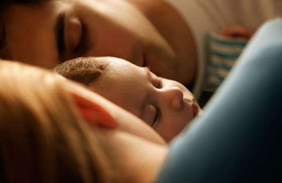 یک زن با موهای بلوند و لباس آبی و یک مرد با موهای تیره و لباس سفید و یک نوزاد بین آنها ، که خوابیده‌اند