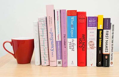 چند کتاب درباره بارداری و فرزندپروری و یک لیوان دسته دار قرمز در کنارشان